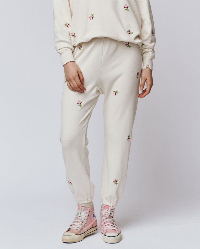 A person wearing THE GREAT. H25 ROSEBUD The Stadium Sweatpant in Washed White With Embroidered Roses, paired with a matching sweatshirt and pink high-top sneakers.