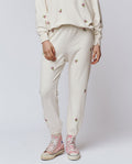 A person wearing THE GREAT. H25 ROSEBUD The Stadium Sweatpant in Washed White With Embroidered Roses, paired with a matching sweatshirt and pink high-top sneakers.