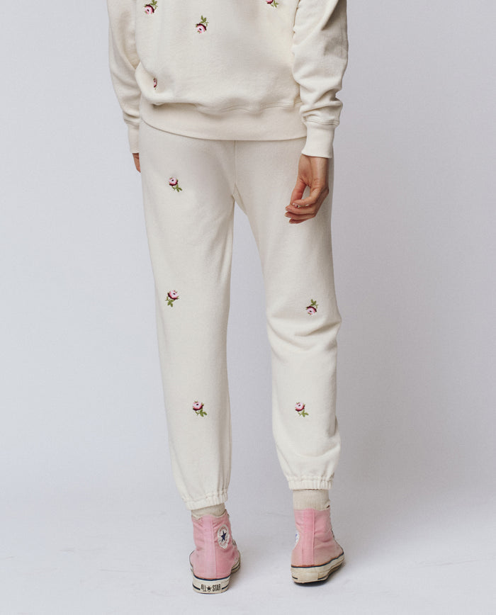 Model wears THE GREAT. H25 ROSEBUD The Stadium Sweatpant in washed white with embroidered roses, styled with a matching sweatshirt and pink high-tops for a vintage athletic look against a plain background.