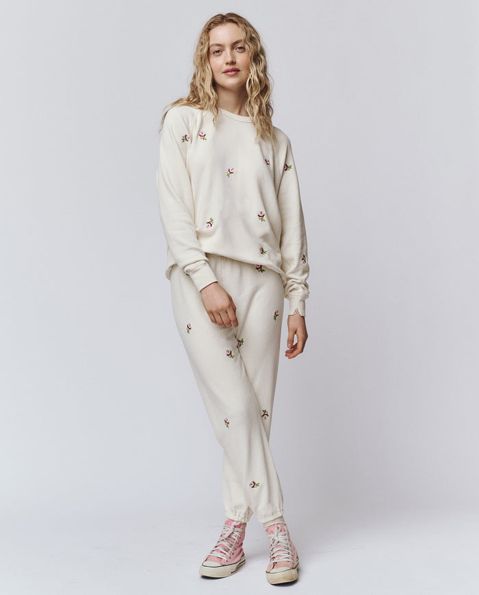 A woman stands against a plain background wearing THE GREAT. H25 ROSEBUD's The Stadium Sweatpant. Embroidered in washed white with rose embroidery, styled with a coordinating sweatshirt and pink high-top sneakers.
