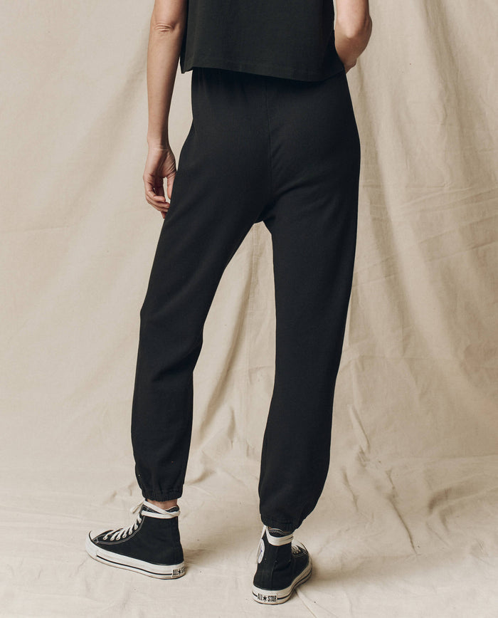 A person in chic black joggers, resembling THE GREAT. CORE KNITS' The Stadium Sweatpant in Almost Black, paired with sneakers, stands on a beige fabric background. These joggers have an elastic waistband for comfort and are made from 100% cotton, providing an ideal mix of style and ease.