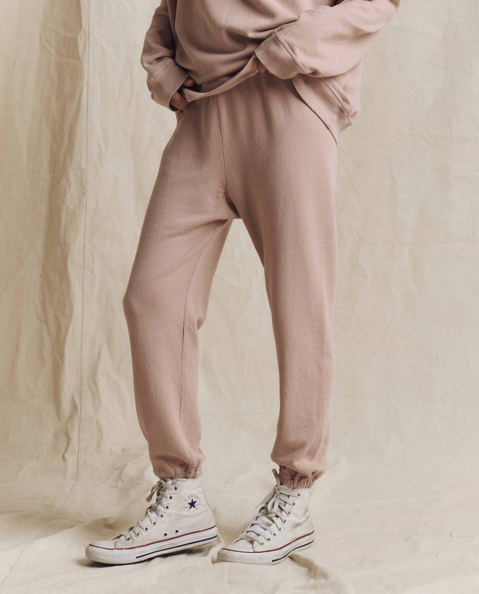 A person wearing THE GREAT. F26 SEASONAL CORE The Stadium Sweatpant. Solid -- Ballet Pink, paired with a matching sweatshirt and white high-top Converse sneakers, stands against a beige fabric backdrop.