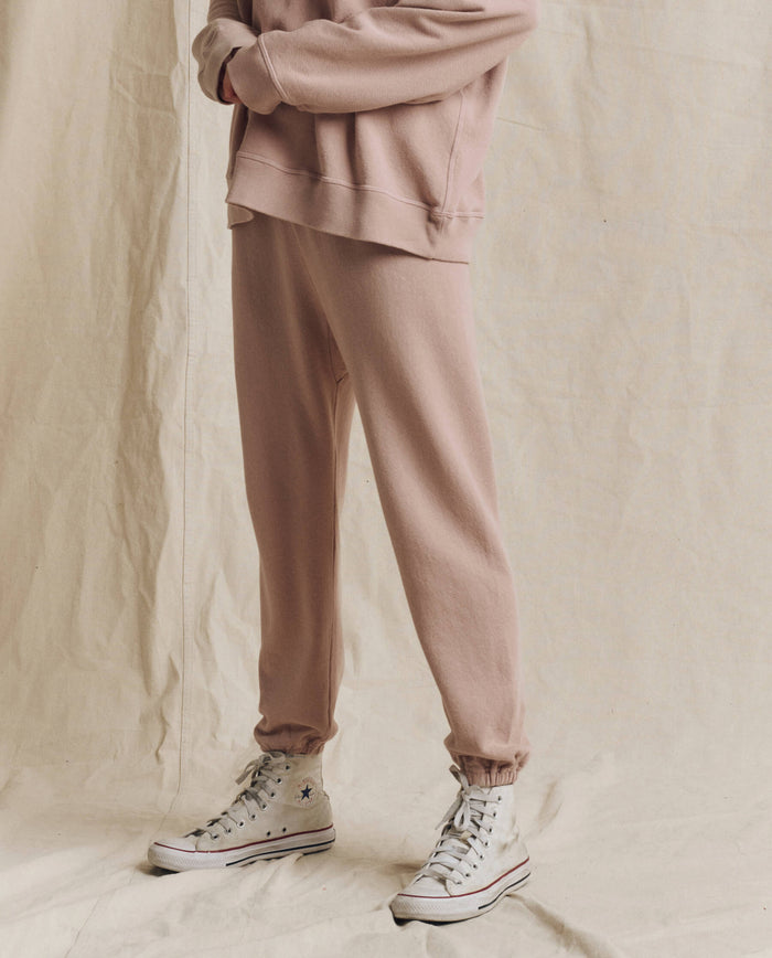 Standing on a beige fabric backdrop in Los Angeles, the person is wearing THE GREAT. F26 SEASONAL CORE’s The Stadium Sweatpant. Solid in Ballet Pink, paired with a light pink sweatshirt and white high-top sneakers. Their face is not visible.