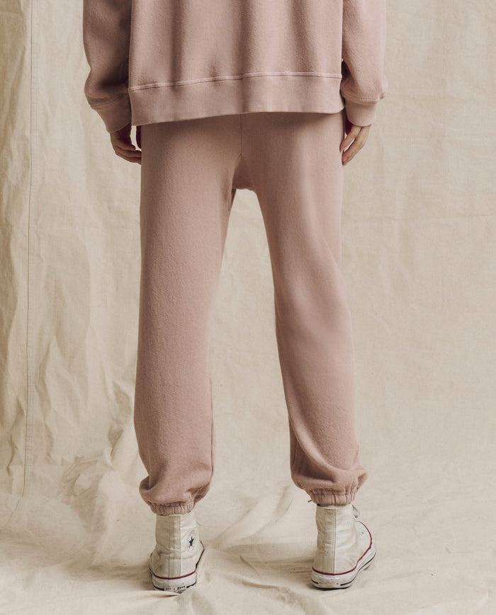 A person stands on a beige fabric backdrop, shown from the back with their head out of frame, wearing THE GREAT. F26 SEASONAL CORE The Stadium Sweatpant in Solid -- Ballet Pink, paired with a matching sweatshirt and white high-top sneakers.