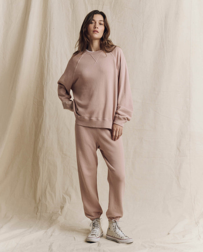 A woman stands against a cream fabric backdrop, wearing THE GREAT. F26 SEASONAL CORE The Stadium Sweatpant in Solid -- Ballet Pink, paired with a matching sweatshirt and white high-top sneakers—a relaxed style inspired by Los Angeles cool.