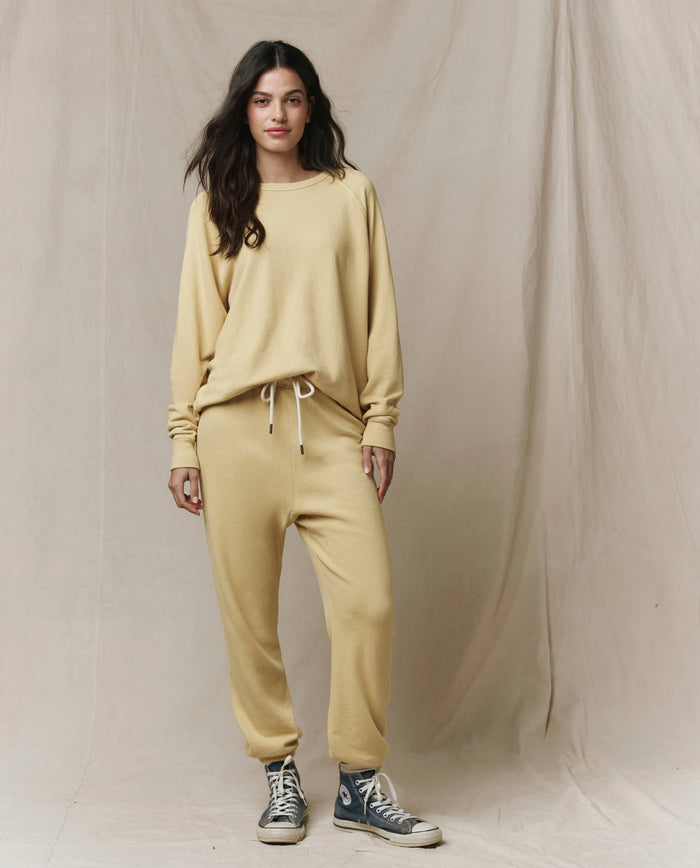 A woman stands before a beige backdrop wearing THE GREAT.'s The Stadium Sweatpant in Butter Yellow, handcrafted for SP26 SEASONAL CORE and inspired by vintage athletic wear, paired with blue high-top sneakers.
