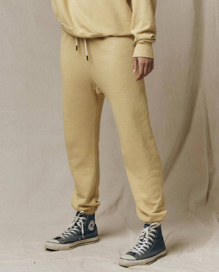 A person in THE GREAT. SP26 SEASONAL CORE The Stadium Sweatpant in Butter Yellow stands against a neutral fabric backdrop, channeling vintage Los Angeles athletic style.