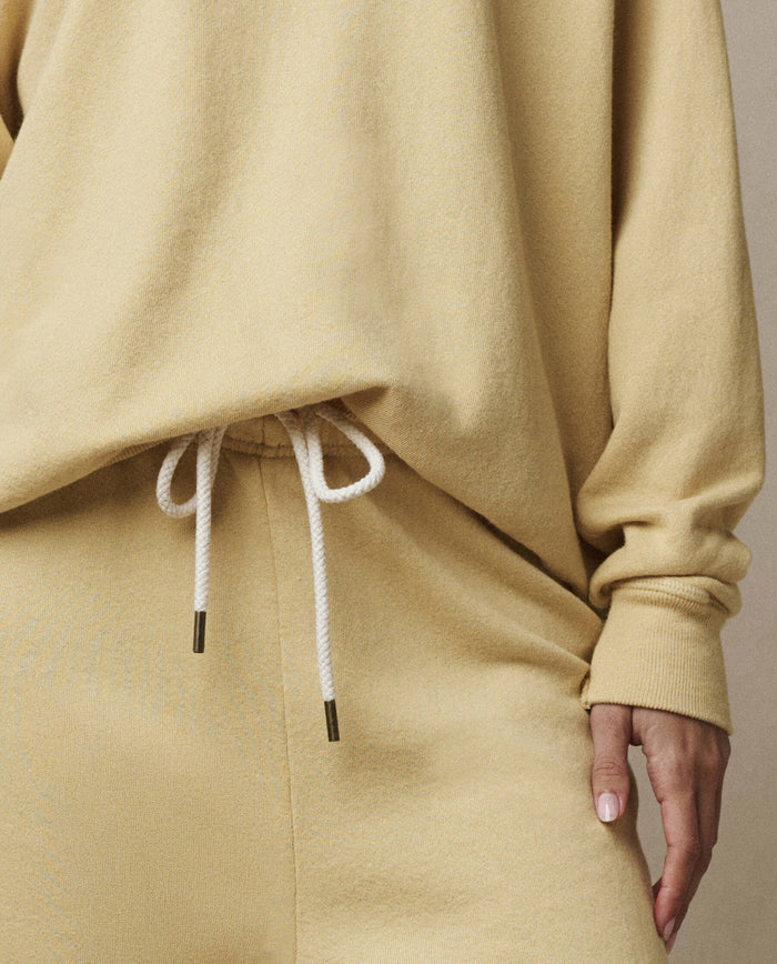 A person wearing a beige long-sleeve top and THE GREAT. SP26 SEASONAL CORE The Stadium Sweatpant in Butter Yellow, featuring white cords and a vintage athletic-inspired design, shown from mid-torso to thigh.
