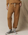Wearing THE GREAT.'s The Stadium Sweatpant in Solid Cork, paired with a matching brown sweatshirt and gray high-top Converse sneakers, the person stands on a beige fabric backdrop.