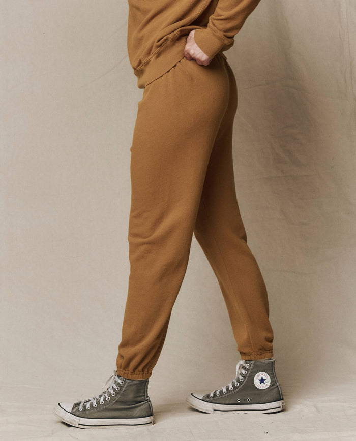 Person wearing The Stadium Sweatpant. Solid -- Cork by THE GREAT. SU26 SEASONAL CORE and gray Converse sneakers, standing sideways against a neutral background.