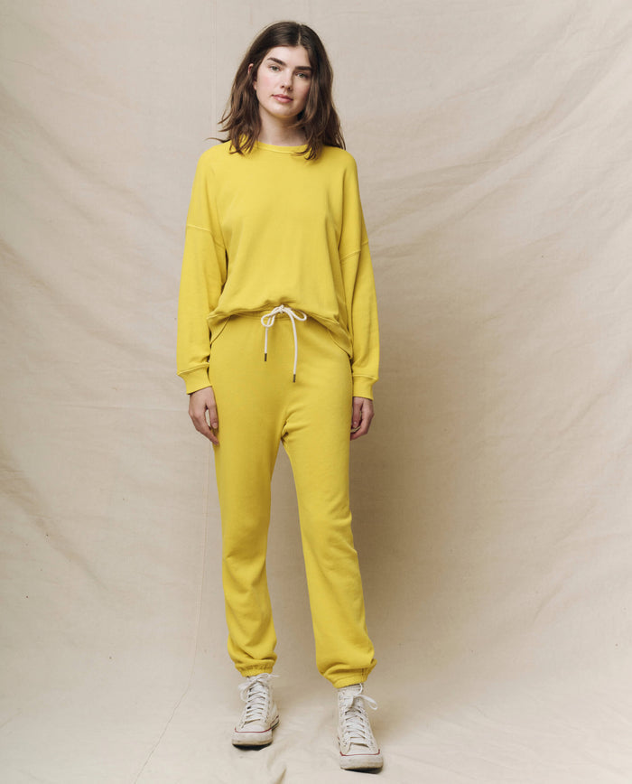 Dressed in The Stadium Sweatpant Solid -- Daffodil by THE GREAT. SP25 KNITS, they stand on a neutral backdrop exuding athleisure vibes with vibrant yellow and crisp white sneakers.