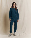 Against a beige backdrop, a person wears THE GREAT. F26 SEASONAL CORE’s The Stadium Sweatpant. Solid in Dark Slate paired with a matching sweatshirt inspired by vintage athletic wear, and gray high-top sneakers.