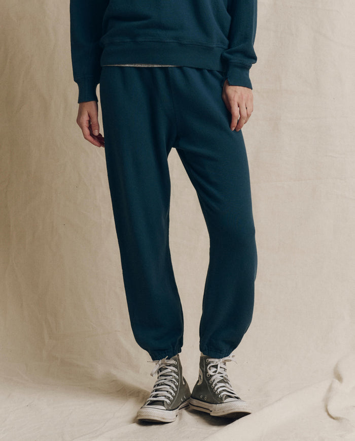 A person wearing a blue sweatshirt, THE GREAT. F26 SEASONAL CORE The Stadium Sweatpant in Solid -- Dark Slate, and grey high-top sneakers stands on a light fabric backdrop, effortlessly channeling vintage athletic wear.