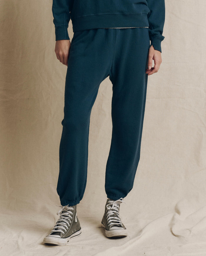 A person stands against a plain beige backdrop wearing THE GREAT. F26 SEASONAL CORE’s The Stadium Sweatpant in Solid -- Dark Slate, paired with a coordinating vintage athletic-inspired sweatshirt and gray high-top sneakers.