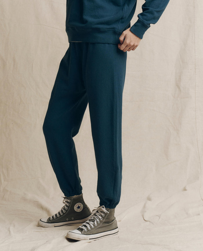 A person stands on a beige fabric background wearing THE GREAT.’s The Stadium Sweatpant in Solid -- Dark Slate, styled with a matching sweatshirt and gray high-top Converse sneakers.