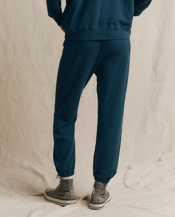 Someone dressed in THE GREAT. F26 SEASONAL CORE The Stadium Sweatpant. Solid -- Dark Slate, styled with a matching sweatshirt and gray high-top sneakers, stands on a neutral fabric backdrop facing away from the camera, capturing an effortless vintage athletic wear vibe.