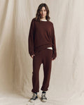 A person stands in front of a beige fabric backdrop, wearing a loose-fitting brown sweatshirt, The Stadium Sweatpant in Driftwood by THE GREAT. F26 SEASONAL CORE—handcrafted for a distinctive vintage feel—and black high-top sneakers.