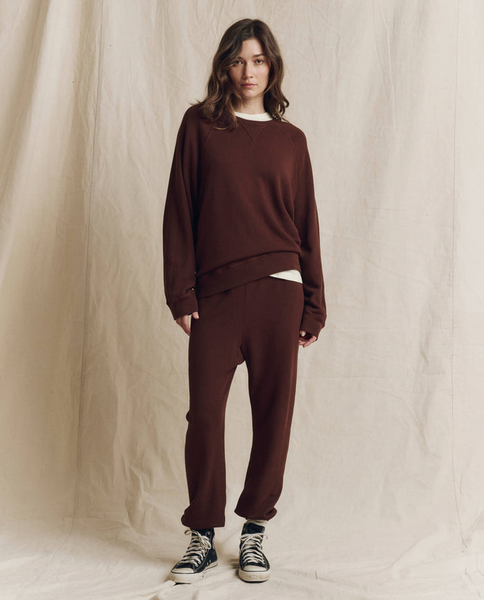 A person stands in front of a beige fabric backdrop, wearing a loose-fitting brown sweatshirt, The Stadium Sweatpant in Driftwood by THE GREAT. F26 SEASONAL CORE—handcrafted for a distinctive vintage feel—and black high-top sneakers.