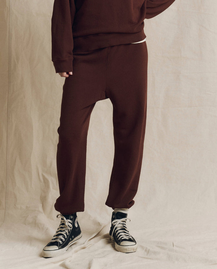 A person wears THE GREAT. F26 SEASONAL CORE’s The Stadium Sweatpant in Solid -- Driftwood, styled with a coordinating brown sweatshirt and black high-top sneakers, standing against a beige fabric backdrop.