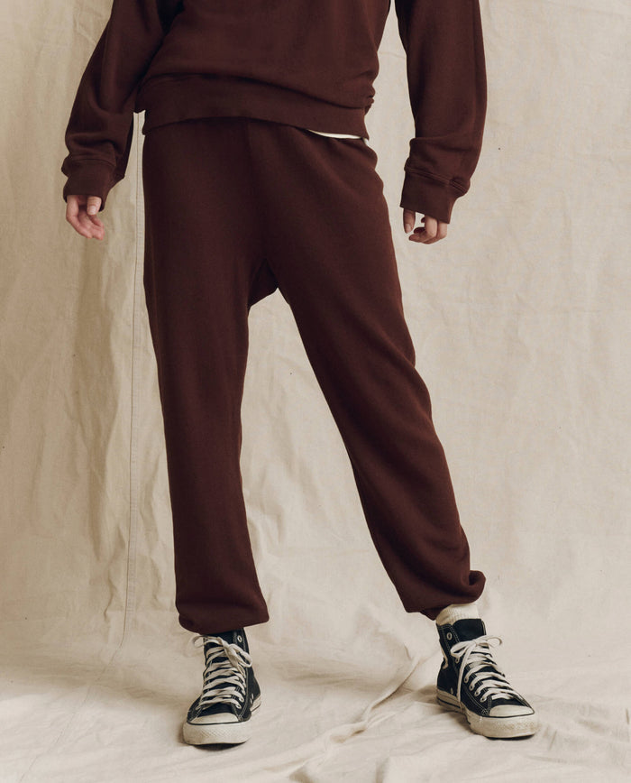 A person stands against a beige backdrop, wearing THE GREAT. F26 SEASONAL CORE The Stadium Sweatpant in Driftwood, paired with a brown sweatshirt and black high-top sneakers. Only the lower half of the body is visible.