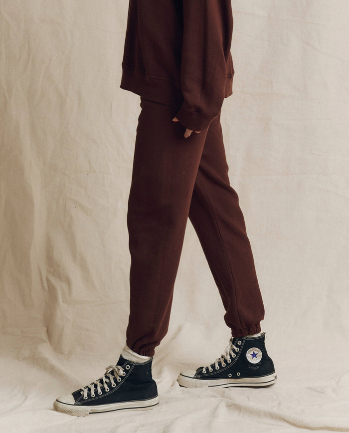 Person wearing THE GREAT. The Stadium Sweatpant. Solid -- Driftwood from the F26 SEASONAL CORE collection, paired with a matching sweater and black high-top Converse sneakers, stands on a light fabric backdrop.
