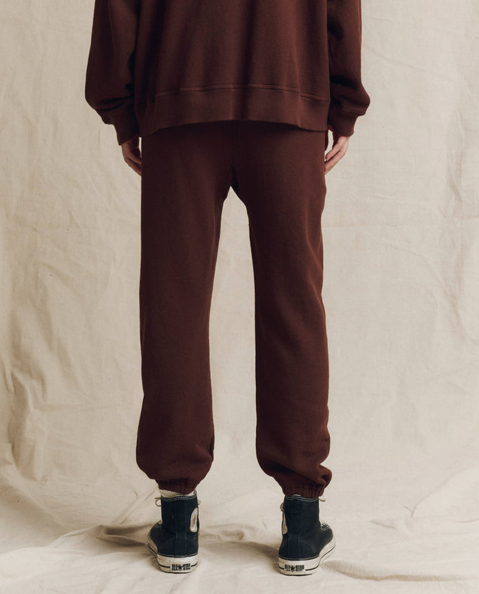 A person stands on a beige fabric background, wearing THE GREAT. F26 SEASONAL CORE Stadium Sweatpant in Driftwood, paired with a brown handcrafted sweatshirt and black high-top sneakers.