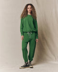 A person with long curly hair wears THE GREAT. SU26 SEASONAL CORE’s The Stadium Sweatpant in Solid Fennel and a matching green sweatshirt with white drawstrings, paired with black high-top sneakers, against a beige fabric backdrop.