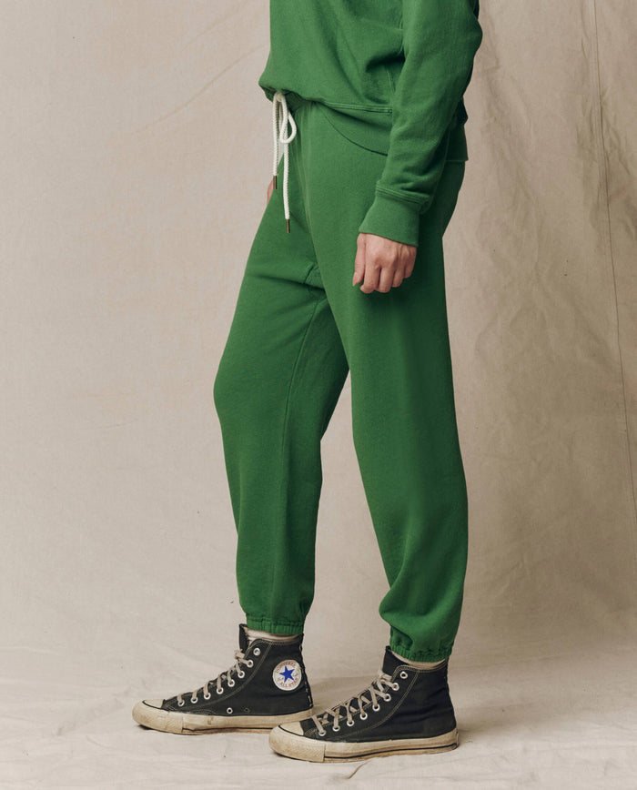 A person stands in The Stadium Sweatpant. Solid -- Fennel by THE GREAT. SU26 SEASONAL CORE, paired with a matching green sweatshirt with white drawstrings and black high-top Converse sneakers, set against a neutral background.