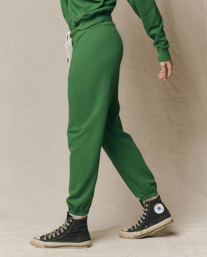 Person wearing The Stadium Sweatpant in Solid Fennel by THE GREAT. SU26 SEASONAL CORE, a matching sweatshirt, and black high-top Converse sneakers, standing sideways against a plain beige background.