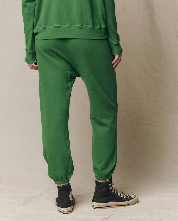A person stands against a plain background, wearing THE GREAT. SU26 SEASONAL CORE The Stadium Sweatpant in Fennel, a matching green sweatshirt, and black high-top sneakers.