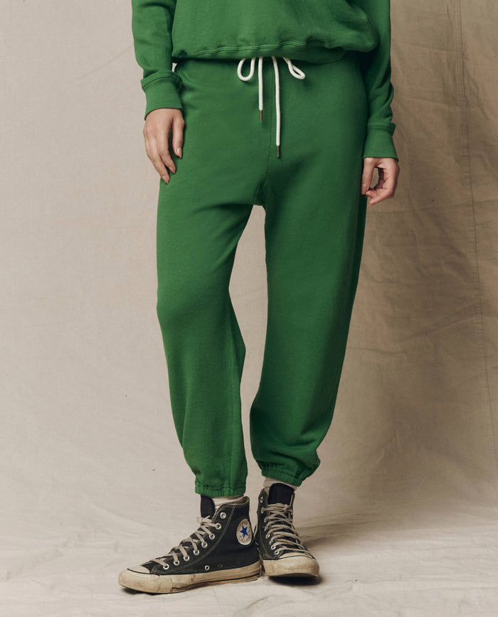 Someone wears The Stadium Sweatpant in Solid Fennel by THE GREAT., styled with a matching green sweatshirt and black Converse sneakers, standing on a neutral backdrop for a fresh take on athletic wear.