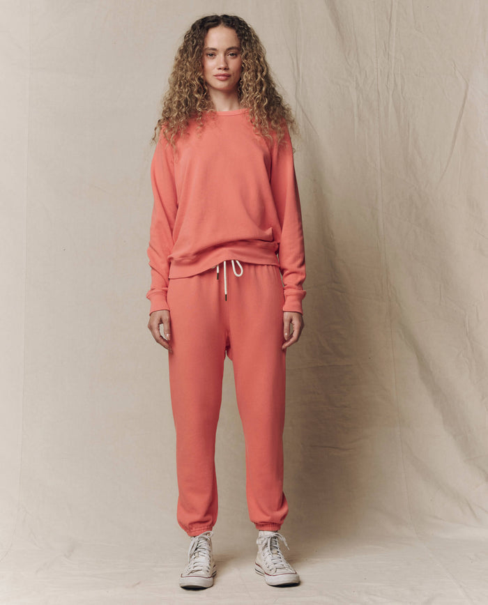 A person faces forward against a neutral fabric backdrop, wearing THE GREAT. The Stadium Sweatpant in Solid Guava (SU26 SEASONAL CORE), paired with a matching sweatshirt and white high-top sneakers.