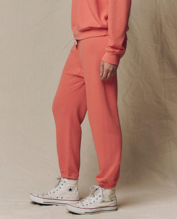 A person in THE GREAT. SU26 SEASONAL CORE’s The Stadium Sweatpant. Solid -- Guava, with a matching sweatshirt and white high-top sneakers, stands against a plain beige backdrop.