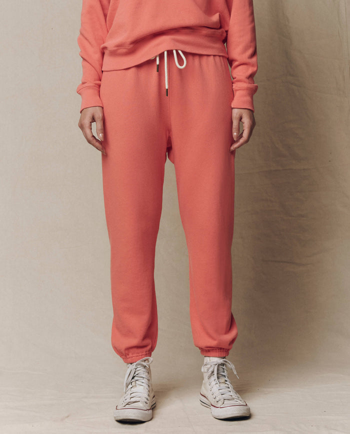 A person in THE GREAT. The Stadium Sweatpant. Solid -- Guava (SU26 SEASONAL CORE) with a matching sweatshirt and white high-top sneakers stands against a neutral background.