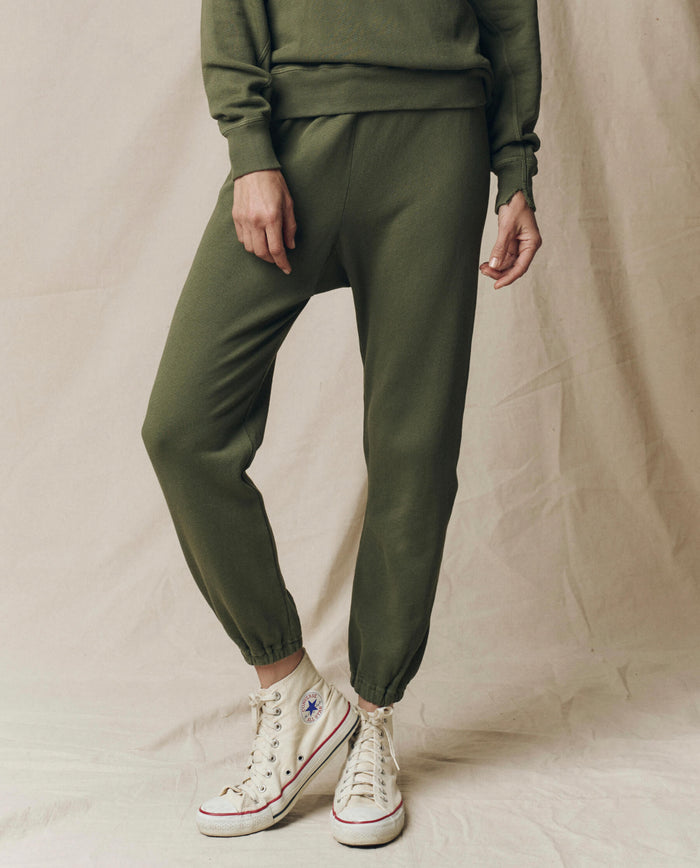 Wearing THE GREAT. SU25 SEASONAL KNITS The Stadium Sweatpant, Solid in Heritage Army, styled with a matching sweatshirt and white high-top Converse sneakers, against a beige fabric backdrop.