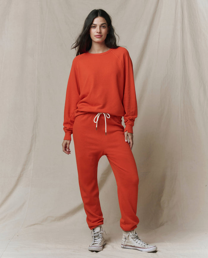 A woman stands against a neutral fabric backdrop wearing THE GREAT. SP26 SEASONAL CORE The Stadium Sweatpant in Persimmon, paired with a matching sweatshirt and white sneakers.