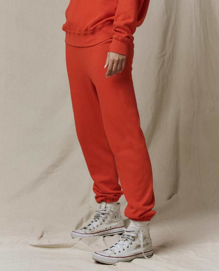 Person wears THE GREAT. SP26 SEASONAL CORE The Stadium Sweatpant in Persimmon with a matching red sweatshirt and white high-top sneakers, standing on a light fabric backdrop.