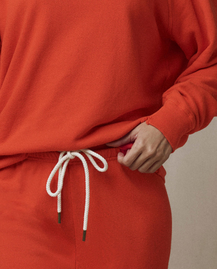 Wearing THE GREAT. SP26 SEASONAL CORE The Stadium Sweatpant in Persimmon, vintage style with a white rope drawstring, hand resting near the waistband.