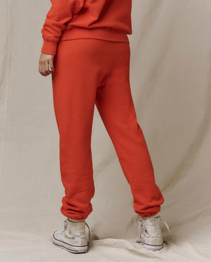 Person stands on a beige fabric backdrop, wearing THE GREAT. SP26 SEASONAL CORE The Stadium Sweatpant in Persimmon with white high-top sneakers—a casual look crafted in Los Angeles.