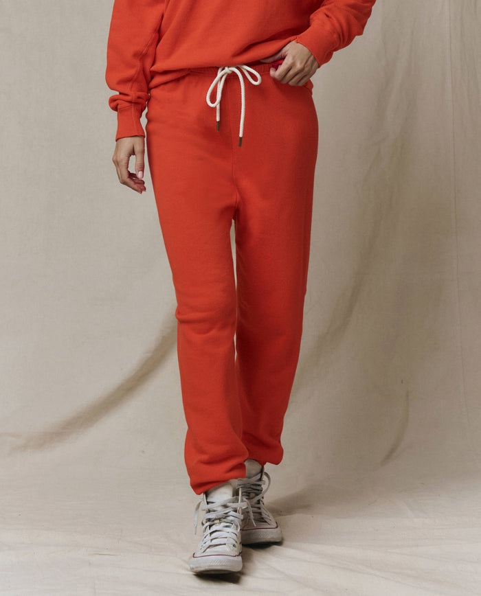A person models THE GREAT. SP26 SEASONAL CORE The Stadium Sweatpant in Persimmon with a matching sweatshirt and white sneakers, set against a plain beige background.