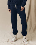 Person wearing The Stadium Sweatpant in True Navy by THE GREAT. FALL 23 KNITS and white sneakers stands on a beige fabric background. The outfit combines the charm of vintage athletic wear with modern comfort.