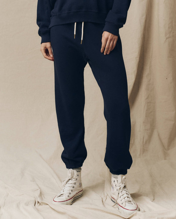 Person wearing The Stadium Sweatpant in True Navy by THE GREAT. FALL 23 KNITS and white sneakers stands on a beige fabric background. The outfit combines the charm of vintage athletic wear with modern comfort.