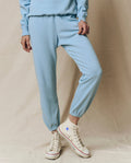 A person wearing THE GREAT. SU25 SEASONAL KNITS The Stadium Sweatpant. Solid -- Vista Blue and matching sweatshirt with white high-top Converse sneakers stands against a beige background.