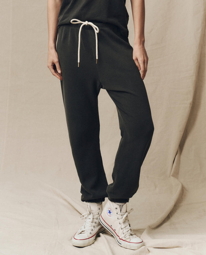 Person wearing The Stadium Sweatpant in washed black from THE GREAT. CORE KNITS with a white drawstring and high-top sneakers, effortlessly blending vintage athletic style against a beige fabric background.