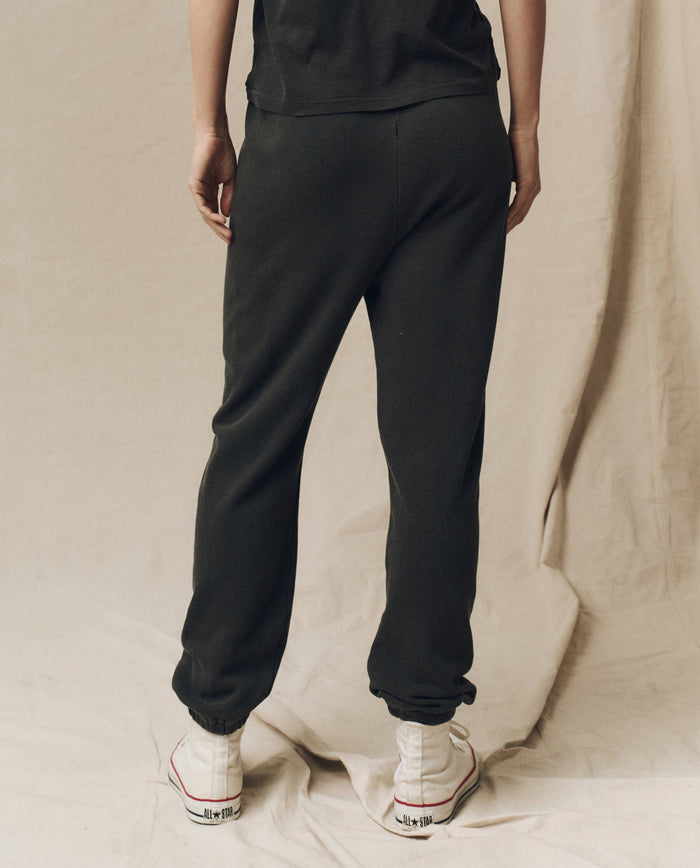 Person standing with their back turned, wearing The Stadium Sweatpant in washed black from THE GREAT. CORE KNITS and white sneakers against a beige background.