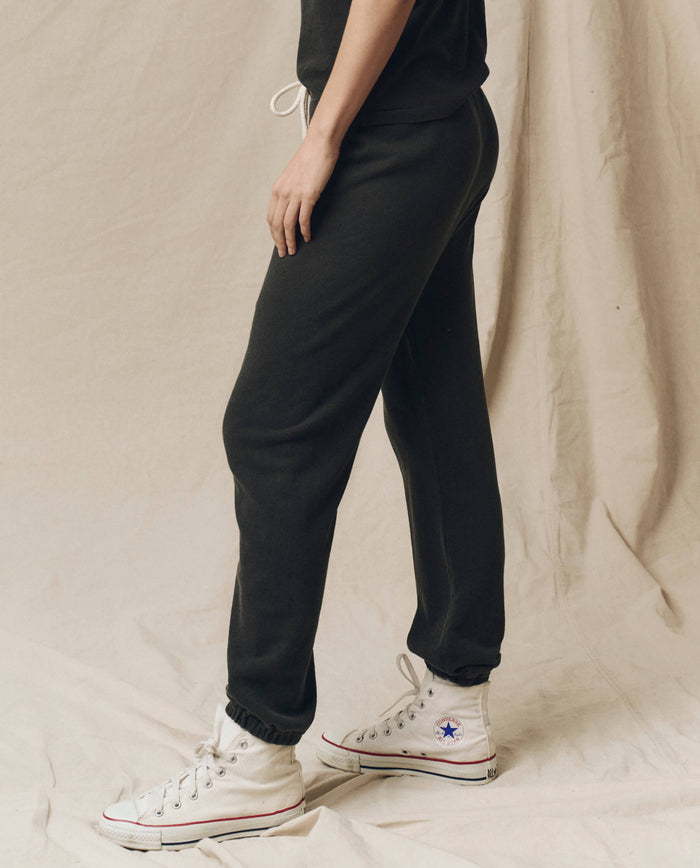 Side view of a person wearing The Stadium Sweatpant in Washed Black from THE GREAT. CORE KNITS and white high-top Converse sneakers, standing on a beige fabric background.