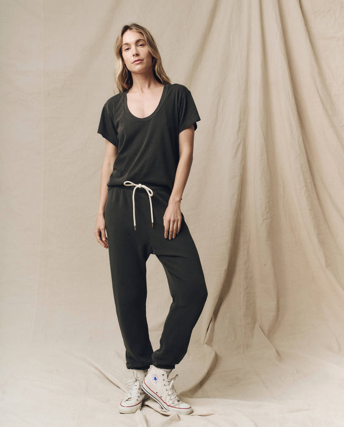 A woman stands against a beige backdrop, wearing The Stadium Sweatpant in washed black from THE GREAT. CORE KNITS and white sneakers, evoking a vintage athletic vibe with its 100% cotton fabric.