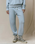 A person wears THE GREAT.’s The Stadium Sweatpant in Whisper Blue from the SP26 Seasonal Core, with matching sweatshirt and off-white high-top sneakers, standing on a neutral background. Inspired by vintage athletic wear, handcrafted in Los Angeles.