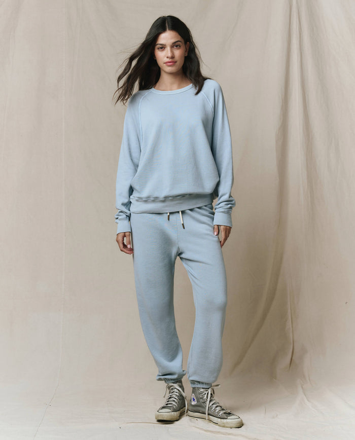 A woman stands before a beige backdrop in THE GREAT.'s SP26 SEASONAL CORE Stadium Sweatpant in Whisper Blue, paired with a matching sweatshirt and gray high-top sneakers—handcrafted in Los Angeles for a vintage athletic look.