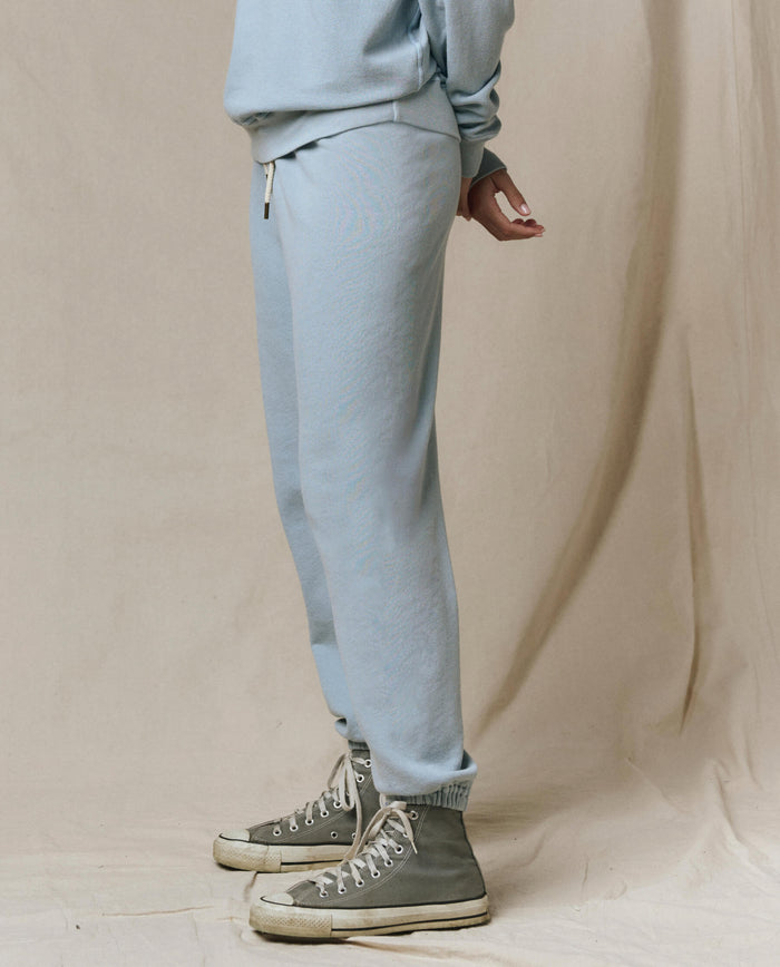 A person stands on a light fabric backdrop wearing vintage athletic wear: THE GREAT. SP26 SEASONAL CORE’s The Stadium Sweatpant in Whisper Blue, paired with a matching sweatshirt and gray high-top sneakers.